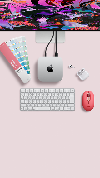 Mac miniを上から見た図。シルバーのアルミニウム製、丸みを帯びた四隅、中央に光沢のあるブラックのAppleのロゴ。コードで接続されている。上部にStudio Displayの一部分が見えている。ケースが開いた状態のAirPods 4、パントンのカラースウォッチ、テンキー付きキーボードとマウス