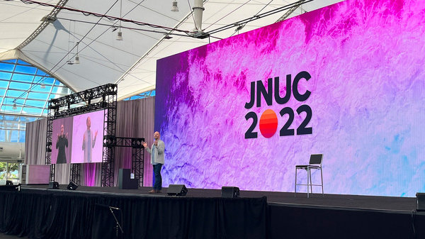 JUNC 2022のロゴが表示された大きなスクリーンを背景にスピーチをしている様子。